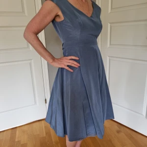 Blå klänning från Phase Eight - Elegant klänning från Phase Eight med v-ringning och knytband under bysten. Sluyar strax nedanför knäna (167 lång). Glansigt tyg med tungt fall, fodrad. Kan knytas både fram och bak. Dragkedja i ryggen. Använd ett tillfälle. Kemtvättad. Storlek 12 (byst 93	midja 76	höft 103).