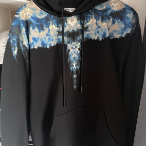 Svart hoodie från Marcelo Burlon  - Säljer en svart hoodie från Marcelo Burlon County of Milan med unikt blått och vitt mönster över axlar och bröst. Säljer då den knappt används och har haft den kanske 2-3 gånger max. Unika med denna tröjan är att den har ett sällsynt mönster och inte alls vanligt att se. Vid frågor är det bara att skriva!