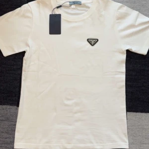 Vit t-shirt från Prada - Säljer en stilren vit t-shirt från Prada med klassisk logga i svart och silver på bröstet. T-shirten har rund hals och korta ärmar, perfekt för en clean och lyxig look.