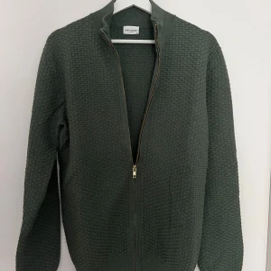 Grön kofta med dragkedja från John Henric - Säljer en stilren grön kofta från John Henric i storlek M. Koftan har ett diskret vävt mönster, hel dragkedja framtill och ribbade muddar vid ärmslut och nederkant. Perfekt för lager-på-lager och enkel att matcha.