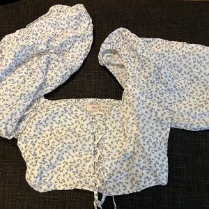 Blus från Nelly - Supersöt blommig croppad blus från Nelly i vitt med små blå blommor. Toppen har långa puffärmar och snörning framtill. inte använda.