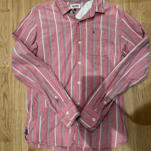 Rosa och vitrandig skjorta från Tommy Hilfiger - Säljer en snygg rosa skjorta med vita och blå ränder från Tommy Hilfiger. Skjortan har lång ärm, klassisk krage och knäppning framtill. Perfekt för dig som vill ha en stilren och fräsch look.