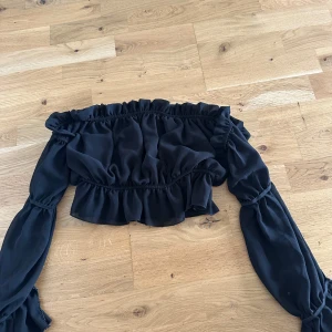 Svart offshoulder blus med volang från Boohoo - Säljer en svart offshoulder blus från Boohoo med långa ärmar och volangdetaljer. Blusen har resår upptill och nertill för en snygg passform och är perfekt för dig som vill ha en trendig och stilren look.