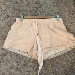 Ljusrosa spets-shorts med knytband - Säljer ett par ljusrosa shorts med spetsdetaljer och knytband i midjan. De har en romantisk känsla med blommigt spetsmönster och mjukt tyg. Perfekta för sommarens varma dagar eller som mysplagg hemma.