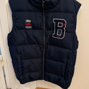 Mörkblå dunväst från Bondelid - Snygg mörkblå dunväst från Bondelid med dragkedja framtill och broderad logga på bröstet. Västen har hög krage och stora fickor på sidorna. Perfekt för lager-på-lager och stilren med sportig känsla. Storlek M.