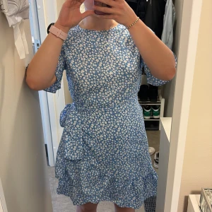 Blå blommig omlottklänning med volang - Söt blå kortklänning med vitt blommönster och volangdetaljer. Klänningen har korta ärmar och knyts i midjan för en fin passform. Perfekt för sommarens alla tillfällen!