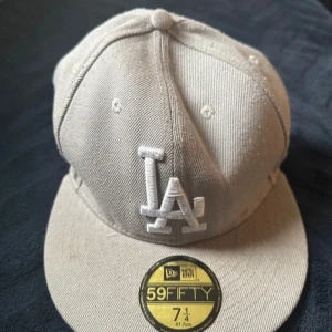 Beige LA-keps från New Era 59FIFTY - Säljer en beige keps från New Era 59FIFTY med LA-logga framtill. Kepsen har en klassisk rak skärm och är i bomullsmaterial. Perfekt för dig som gillar sportiga accessoarer och vill ha en stilren look.