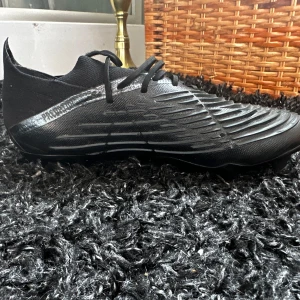 Svarta Adidas predictor edge.1low - Säljer ett par svarta Adidas fotbollsskor med blå och röda ränder på sidan. Skorna har snörning, låg profil och mönstrad ovansida för bättre bollkontroll. Sulan har rejäla dobbar för bra grepp på planen.