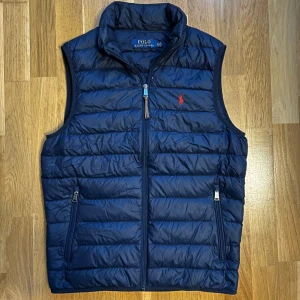Mörkblå dunväst från Polo Ralph Lauren - Snygg mörkblå dunväst från Polo Ralph Lauren med ståkrage och dragkedja framtill. Västen har två fickor med dragkedja och klassisk röd logga broderad på bröstet.