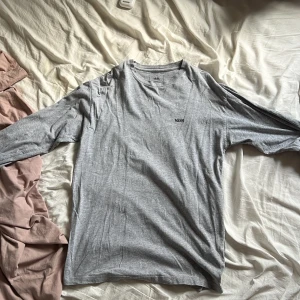 Grå långärmad tröja från Vans - Säljer en grå långärmad tröja från Vans i klassisk passform. Tröjan är i mjuk bomullsmix och har en liten Vans-logga tryckt på bröstet. Perfekt för en avslappnad stil.