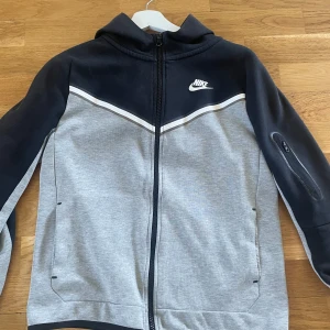 Nike ziphoodie i svart och grå - Nike ziphoodie i svart och ljusgrå med vit logga på bröstet. Jackan har dragkedja framtill, huva och en extra ficka med dragkedja på ärmen. Storlek 158-170 Cm! 
