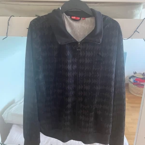 Svart mönstrad jacka från Puma - Säljer en svart jacka från Puma med diskret geometriskt mönster. Jackan har dragkedja framtill, hög krage och ribbade muddar vid ärmslut och nederkant. Perfekt för dig som gillar sportig stil och vill ha något bekvämt och snyggt.