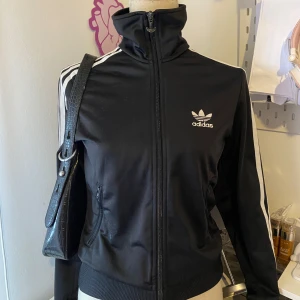 Svart Adidas track jacket med vita ränder - Klassisk svart Adidas track jacket med vita ränder längs ärmarna och vit Adidas-logga på bröstet och ryggen. Jackan har dragkedja framtill, hög krage och ribbade muddar. Perfekt för en sportig och avslappnad stil.