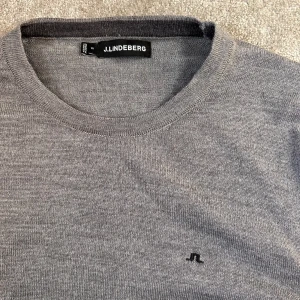 Grå stickad tröja från J.Lindeberg - Säljer en stilren grå stickad tröja från J.Lindeberg med rund halsringning och diskret logga på bröstet. Perfekt för lager-på-lager och passar till många olika outfits.