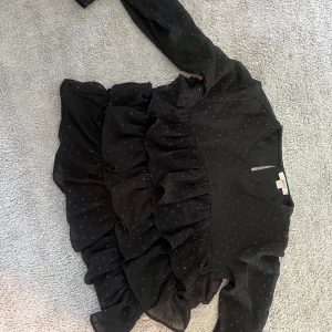 Svart volangblus från Gina Tricot - Supersöt svart blus från Gina Tricot med små diskreta prickar och flera lager volanger framtill. Långärmad modell med lätt transparent tyg och rund halsringning. Perfekt för dig som gillar en trendig och lekfull stil!