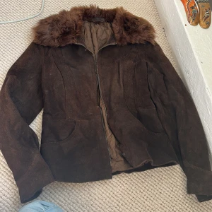 Brun mockajacka med fuskpäls - Säljer en brun mockajacka med mjuk fuskpäls runt kragen. Jackan har dragkedja framtill, två fickor och quiltat foder på insidan. Perfekt för kyligare dagar och ger en snygg vintagekänsla.