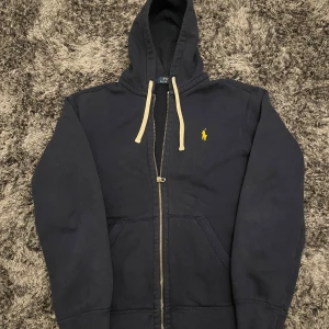 Marinblå hoodie från Polo Ralph Lauren - Snygg marinblå hoodie från Polo Ralph Lauren med dragkedja och klassisk gul broderad logga på bröstet. Tröjan har huva med vita snören och känguruficka framtill. Tillverkad i mjuk bomull, perfekt för chill dagar eller när du vill ha en clean look.
