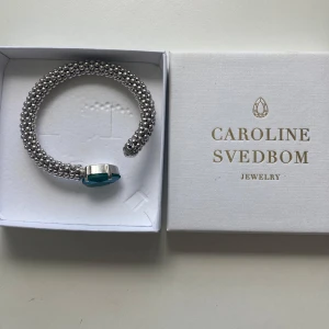 Silverarmband Caroline Svedbom med blå sten - Snyggt armband från Caroline Svedbom i silverfärg med pärlstruktur och en stor, droppformad blågrön sten framtill. Armbandet är stelt och öppet i modellen, vilket gör det lätt att ta på och av. Perfekt statement piece för att lyfta din outfit.
