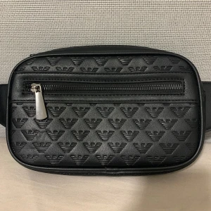 Svart magväska från Emporio Armani - Snygg svart magväska från Emporio Armani med präglade loggor över hela väskan. Väskan har justerbart band, dragkedja framtill och flera fack inuti. Tillverkad i skinnimitation med stilren look och metall-dragkedja framtill.