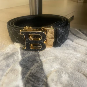 Burrbery!!Svart bälte med guldspänne och B-mönster - Snyggt svart burrbery bälte med ett stort guldigt B-spänne. Bältet har ett diskret mönster med små B:n över hela bandet. Perfekt accessoar för att lyfta din outfit med en lyxig touch.