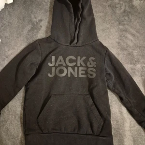 Svart hoodie från Jack & Jones - Svart hoodie från Jack & Jones med stor logga framtill. Klassisk känguruficka och huva. Tillverkad i mjuk bomullsblandning, perfekt för chill eller skola. Långärmad och normal passform.