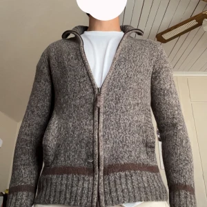 Brunmelerad kofta från Marwin,  - Säljer en brunmelerad stickad kofta från Marwin i storlek S men sitter lite mer som XS. Koftan har dragkedja framtill, hög krage och två fickor. Den är ribbstickad vid ärmslut och nederkant samt har en mörkbrun rand längs midja och ärmar. Perfekt för kyliga dagar.