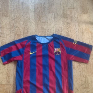 Barcelona Ronaldinho 10 fotbollströja - Klassisk FC Barcelona fotbollströja från Nike med Ronaldinho och nummer 10 på ryggen. Tröjan har breda blå och röda ränder, klubbmärke på bröstet och gul Nike-logga. Tillverkad i lätt och ventilerande material, perfekt för match eller träning.