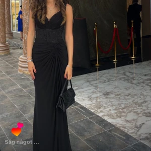 Elegant svart långklänning - Supersnygg svart långklänning med v-ringning och draperad midja. Klänningen har mellan smala axelband och markerad midja med fina detaljer. Perfekt för bal eller fest, ger en riktigt classy look. Orginalpris 3000, endast använd en gång 