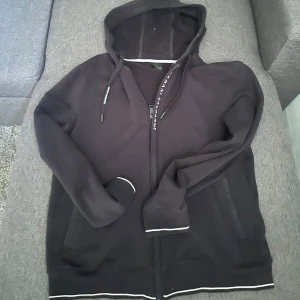 Svart hoodie från Armani Exchange - Svart hoodie från Armani Exchange med dragkedja och huva. Snygga vita detaljer vid muddar och nederkant samt logga på snörena. Perfekt för en chill och stilren look. Långärmad och i mjuk bomull.