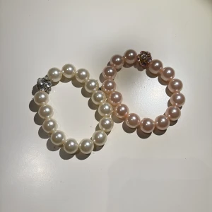 Två pärlarmband med glittriga detaljer - Säljer två elastiska armband med stora pärlor, ett i vitt och ett i ljusrosa. Båda har en glittrig blomformad detalj med stenar som ger extra bling. Perfekta för att lyfta din outfit med en söt och elegant touch.