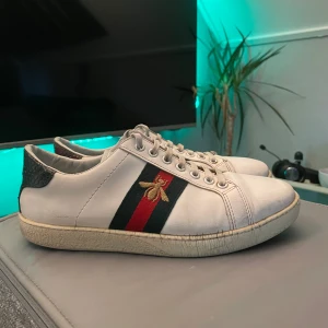 Gucci Ace vita sneakers med bi-detalj - Gucci Ace sneakers i vitt skinn med klassiska gröna och röda ränder på sidan och broderad guldfärgad bi. Hälkappan har ormskinnsmönster i rött och grönt. Platt sula och snörning framtill. Perfekta för dig som vill sticka ut med lyxig streetstyle.