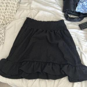 Svart volangkjol med resårmidja - Supersöt svart kortkjol med bred resår i midjan och volang längst ner. Kjolen har en lätt strukturerad yta och är perfekt för dig som gillar en enkel men ändå lekfull stil.