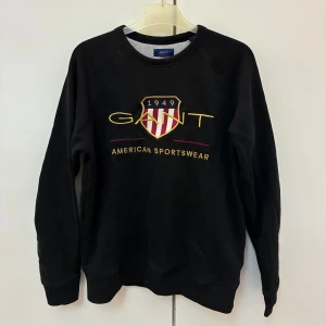 Svart sweatshirt från GANT med brodyr - Svart sweatshirt från GANT med broderad logga och sköld på bröstet i gult, rött och vitt. Klassisk rund halsringning och långa ärmar. Mjuk insida och ribbade muddar vid ärmslut och nederkant. Sitter som Storlek S/XS. Köpt för 800kr. 