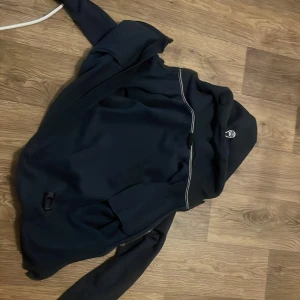 Svart softshelljacka Sail Racing - Svart softshelljacka från Sail Racing med huva och dragkedja. Jackan har logga på bröstet och ärmarna, samt praktiska fickor med dragkedja. Perfekt för dig som gillar stilren och sportig look. Materialet känns mjukt och följsamt.