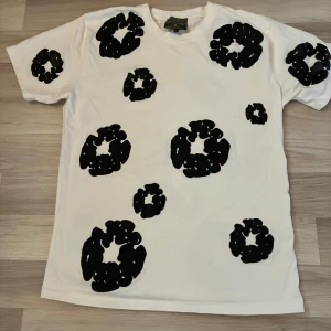Vit t-shirt med svart mönster Denim Tears - Säljer en vit t-shirt från Denim Tears i storlek XXL men är M. T-shirten har coola svarta mönster över hela plagget och klassisk passform med korta ärmar. Materialet är mjuk bomull som känns skönt mot huden. Perfekt för dig som gillar streetwear med unik vibe.