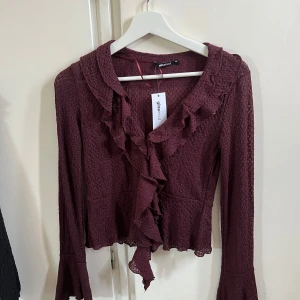 Vinröd volangblus från Gina Tricot - Supersöt vinröd blus från Gina Tricot med volangdetaljer längs krage och framtill. Långärmad modell i lätt strukturerat tyg, perfekt för dig som gillar romantiska vibes. Blusen har en feminin v-ringning och snygg passform. Helt ny