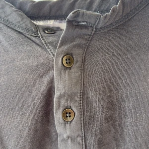 Mörkgrå t-shirt med knappar från Zara - Snygg mörkgrå t-shirt från Zara med korta ärmar och tre knappar vid halsen. Tillverkad i mjuk bomull och har en avslappnad passform. Perfekt för en chill och stilren look.