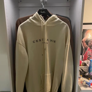 Beige hoodie från  C'est La Vie - Säljer en beige hoodie med svart text 'C'est La Vie Paris' på bröstet. Tröjan har huva med snörning och en stor magficka. Perfekt för dig som gillar en clean och chill stil. Storlek m. Aldrig använd