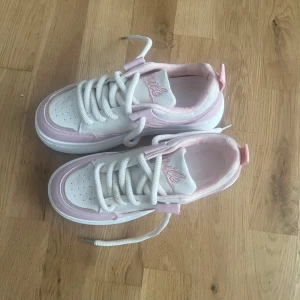 Vita och rosa sneakers med snörning - Snygga sneakers i vitt och ljusrosa med tjocka vita snören och broderad text på plösen. Skorna har rund tå, perforerade detaljer framtill och en chunky sula. Perfekta för barn och färgglada festligheter.