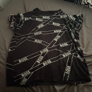 Svart BOSS t-shirt med logomönster - Svart t-shirt från BOSS med coolt vitt logomönster över hela tröjan. Klassisk passform och korta ärmar, perfekt för dig som gillar streetstyle och vill sticka ut. Materialet känns mjukt och skönt mot huden.