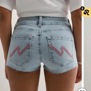 Ljusblå jeansshorts med broderi - Snygga ljusblå jeansshorts med rå kant och coola bakfickor med rosa broderad zigzag-detalj. Hög midja och klassisk femficksmodell. Perfekta för varma dagar och en avslappnad look. Säljer dessa pågrund av att jag köpte en för stor storlek, dessa som jag säljer är i storlek 38 och vid fler frågor är det bara att kontakta mig. Dem är helt nya bara testat dem