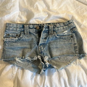 Ljusblå jeansshorts med fransig kant - Snygga ljusblå jeansshorts med fransig och sliten kant, klassisk femficksmodell och bälteshällor. Shortsen har råa kanter och slitningar framtill för en cool, avslappnad vibe. Perfekta för varma dagar och festivalhäng. OBS! Kan skicka som tidigast 8e augusti för är bortrest tills dess
