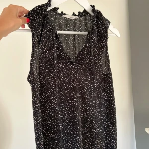 Svart prickig blus från Mango - Supersöt svart blus från Mango med rosa prickar och volangdetaljer vid kragen. Ärmlös modell med knytband vid halsen💕💕🌸🌸