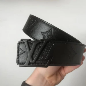 Svart Louis Vuitton skärp i skinn - Snyggt svart skärp från Louis Vuitton i skinn med präglad LV-logga och monogramdetaljer längs hela bältet. Bältet har en bred design och ett stilrent LV-spänne i matt svart. Perfekt för att lyfta vilken outfit som helst.