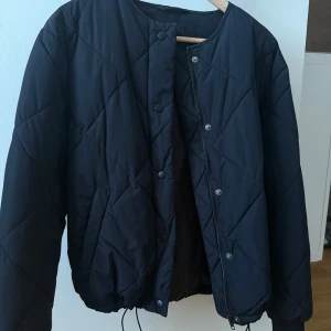 Svart quiltad pufferjacka - Riktigt snygg & trendig svart pufferjacka med quiltat mönster och rund halsringning. Jackan har dragkedja och tryckknappar framtill samt snörning i nederkant för extra passform. Perfekt för kyliga dagar och enkel att styla till olika outfits. Använd 1 gång