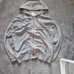 Grå hoodie från HOPE Stockholm - Grå hoodie från HOPE Stockholm med dragkedja, huva och vita snören. Tillverkad i 100% bomull och har klassiska kängurufickor framtill. Perfekt för en chill och avslappnad stil.