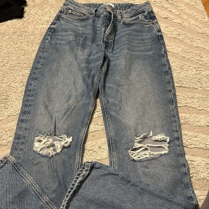 Blå slitna jeans med hål på knäna - Säljer ett par blå jeans med snygga slitningar och hål på båda knäna. Jeansen har klassisk femficksdesign, normal passform och är tillverkade i jeansmaterial. Perfekta för en avslappnad och trendig look.