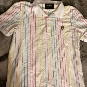 Randig pikétröja från Lyle & Scott - Vit pikétröja från Lyle & Scott med smala ränder i rött, gult och grönt. Klassisk krage, korta ärmar och knäppning framtill. Broderad gul logga på bröstet. Är en storlek XL men är mycket liten i storlek.