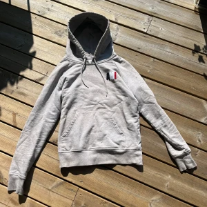 Grå hoodie från Ami Paris  - Snygg grå hoodie från Ami Paris i mjuk bomull. Tröjan har huva med snörning, känguruficka och broderad logga i mörkblått , vitt och rött på bröstet. Perfekt för en chill och stilren look.