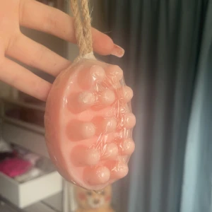 Rosa massage-tvål med snöre - En rosa tvål med massageeffekt, formad med rundade knoppar för extra skön känsla. Kommer med ett praktiskt snöre för enkel upphängning i duschen. Tvålen är inslagen i plast och har en mjuk, pastellig färg. Perfekt för en lyxig duschrutin.